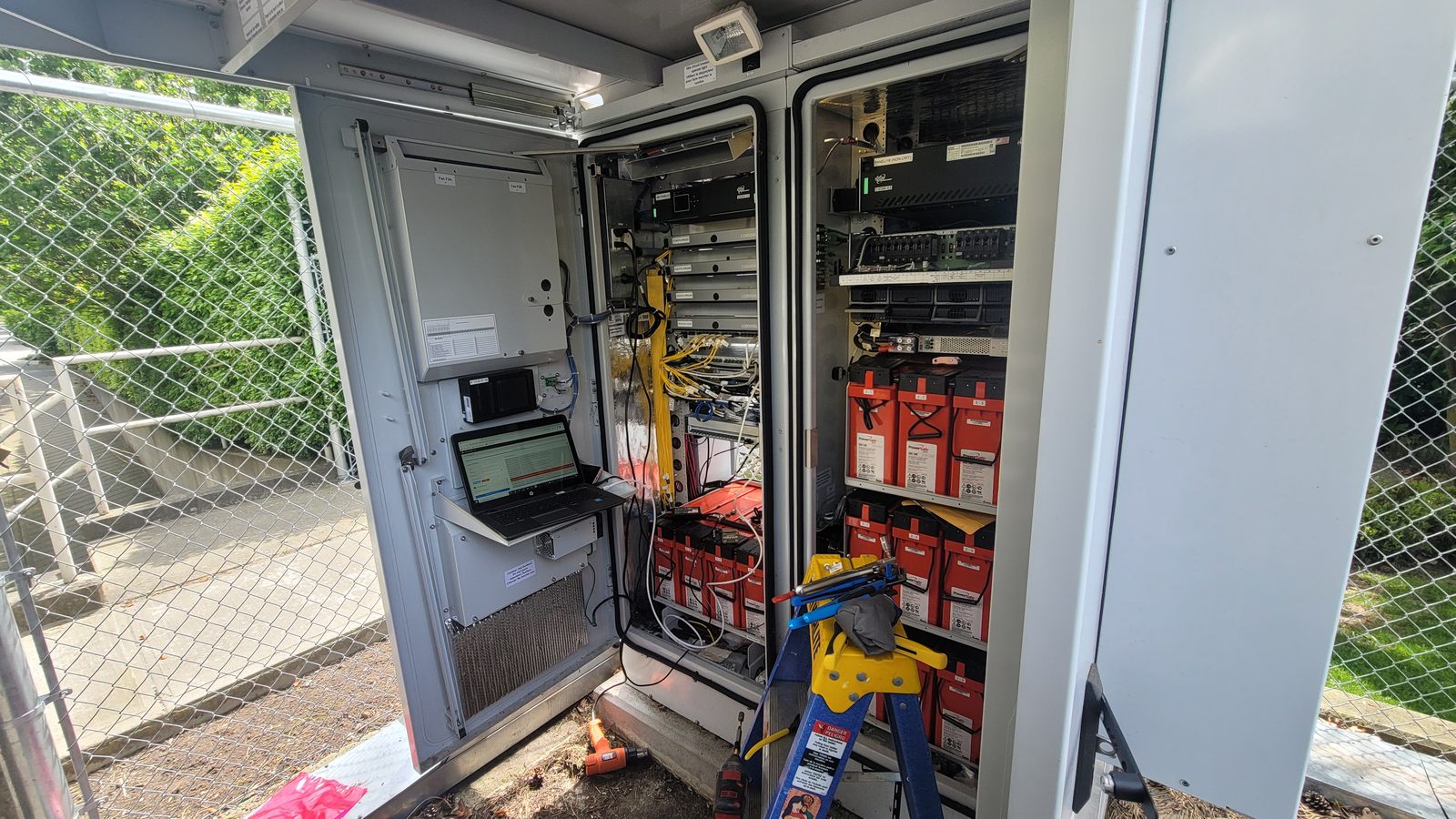 Telecom equipment cabinet with battery bank, fiber links, and radio gear being serviced